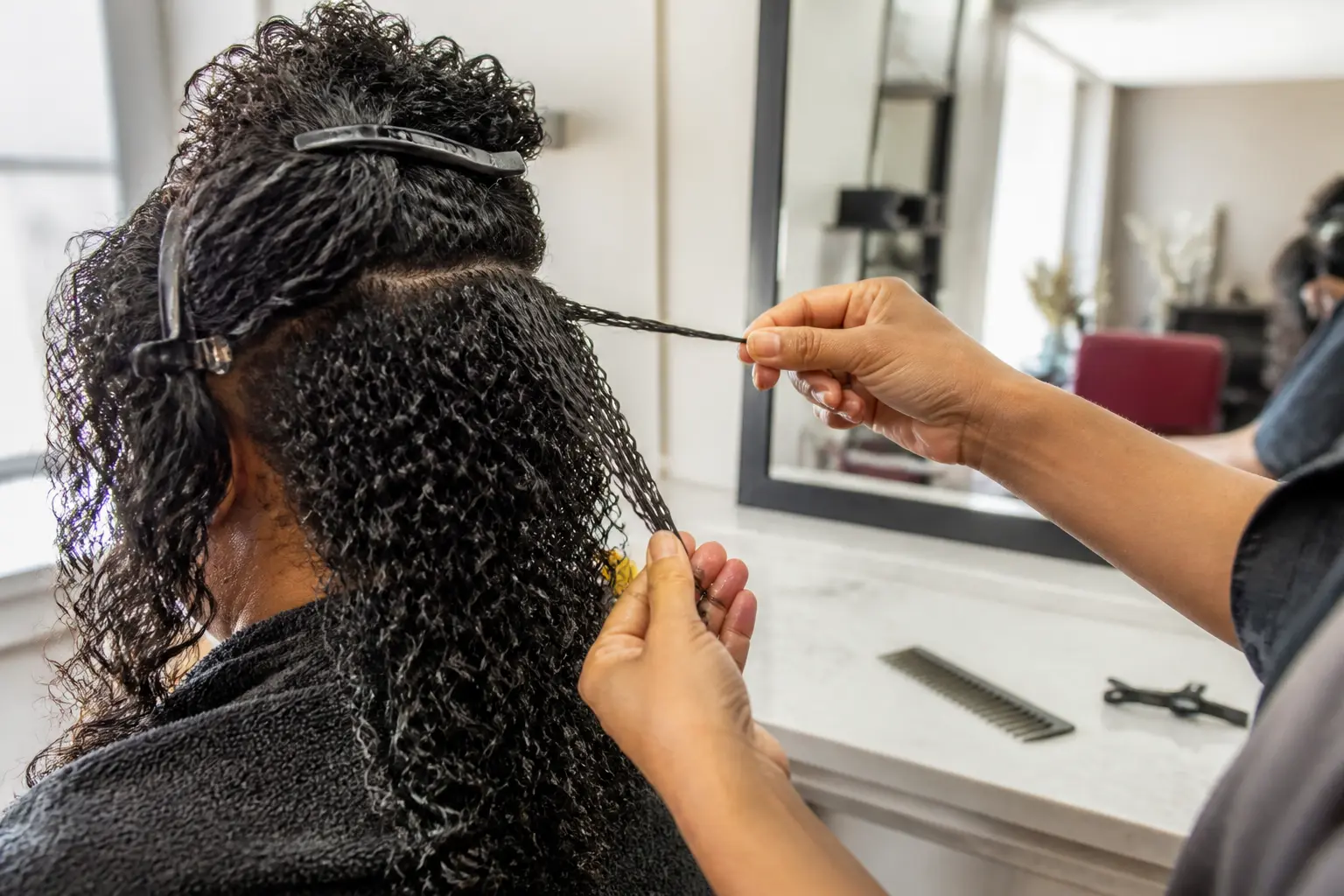 Stylistes couper les cheveux frisés en observant une boucle humide étirée dans un salon lumineux, miroir en fond