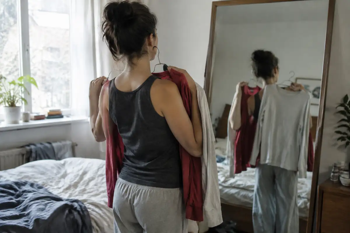 Une femme de dos devant un miroir compare un haut rouge et un haut gris