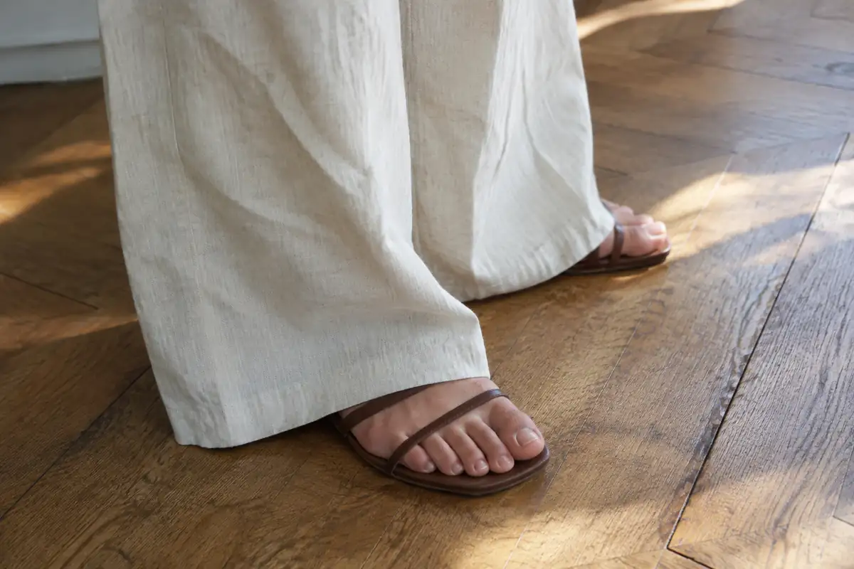 Pantalon large en lin écru qui retombe sur une sandale plate à brides fines, sur un parquet en chêne
